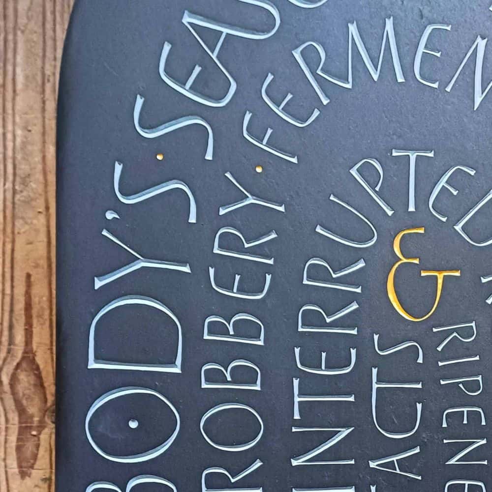 a close up of carved lettering in curved edge of slate, visible words include body's seasons, robbery, ferment, interrupted, and an ampersand in gold